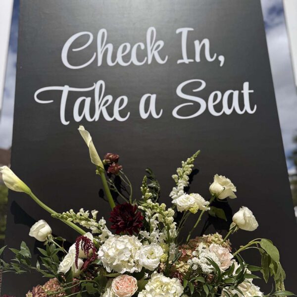 “Check In, Take a Seat” sign on black panel with elegant white, blush, and burgundy floral arrangement.