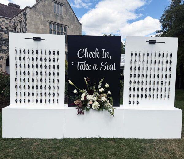 Large black and white wedding seating chart wall with modern key tags and fresh floral arrangement outdoors.