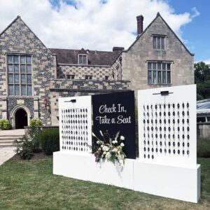 Elegant black and white wedding seating chart wall with floral arrangement reading “Check In, Take a Seat,” displayed outside a historic venue.